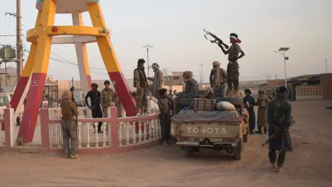 Des combattants en tenue camouflage autour d'un pick-up sur une route poussiéreuse au Mali. Un homme en silhouette se tient sur le véhicule avec un fusil automatique. À gauche, un monument aux jambes jaunes et brunes est visible.